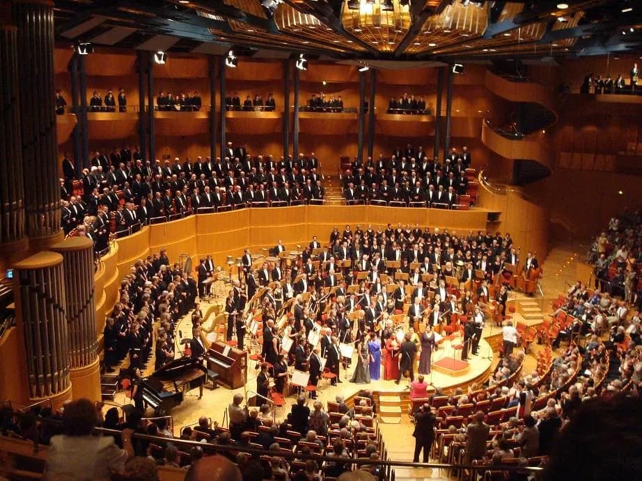 A large orchestra and choir performing on stage in a grand concert hall filled with an audience.