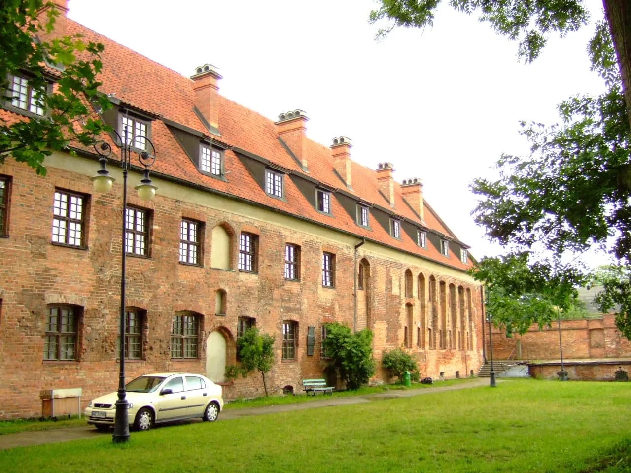 A long, red-brick building with a red-tiled roof and multiple dormer windows, with a light-colored car parked on a paved path in front of it.